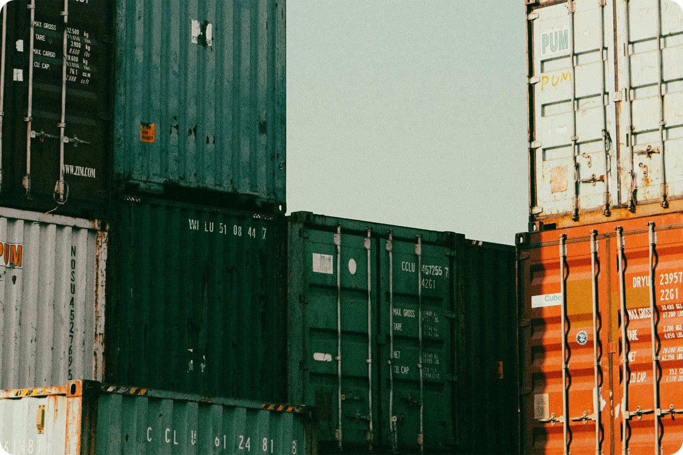 Stacked shipping containers in green, white, and orange at a port, showing cargo weight and identification markings