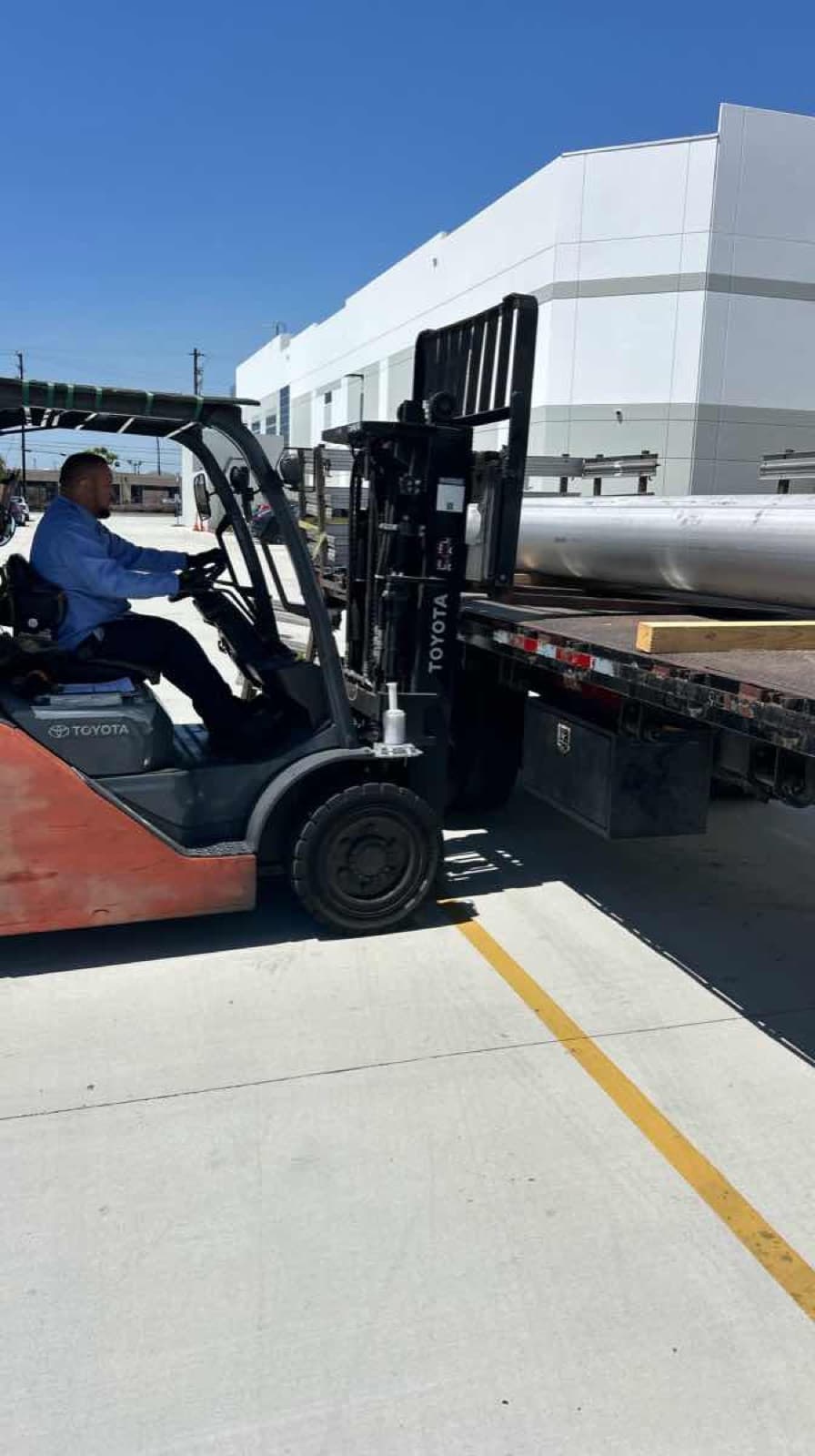 Orange Toyota forklift loading bundles of aluminum cylindrical tubing onto a Curri flatbed truck in a sunny industrial yard, ready for a same-day Moffett-equipped delivery