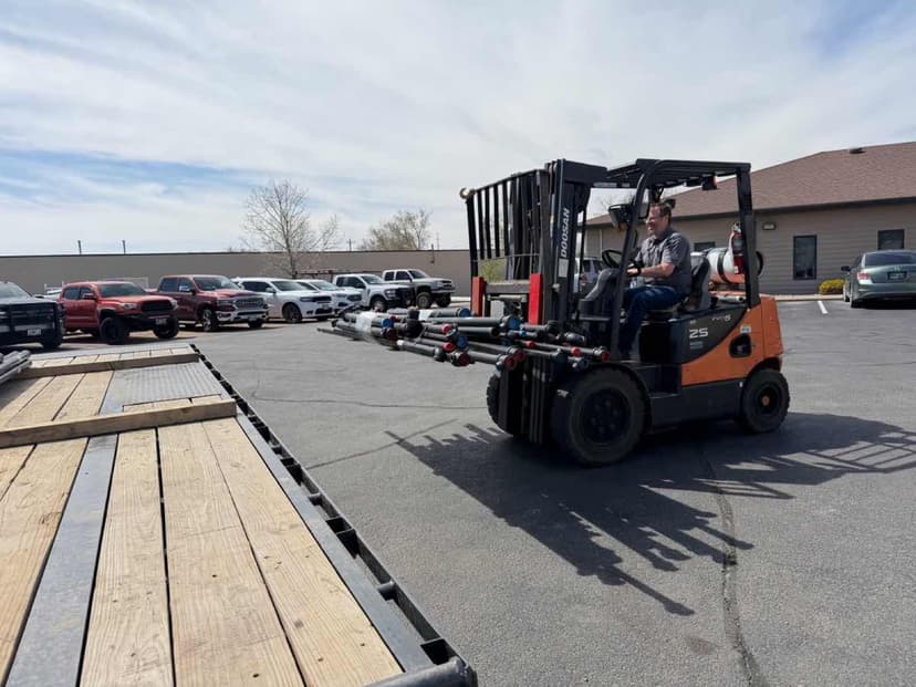 Orange Doosan forklift carrying long pipes near a Curri flatbed for an industrial-supply drop-off