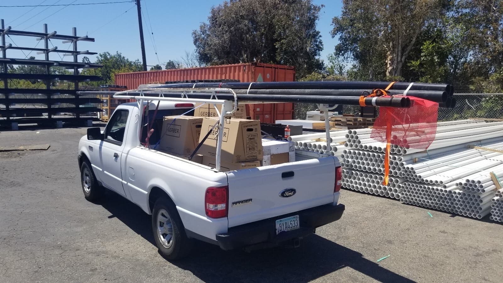 White Ford Ranger pickup with overhead pipe rack hauling long black pipe at a Curri supply yard pickup