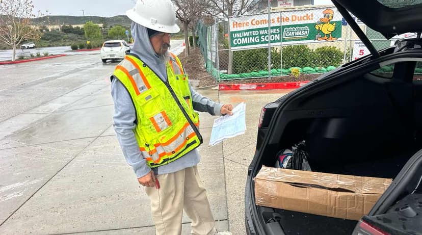 Worker in a hi-vis vest reviewing plans next to an open SUV trunk with cardboard cargo on a Curri delivery-site drop-off