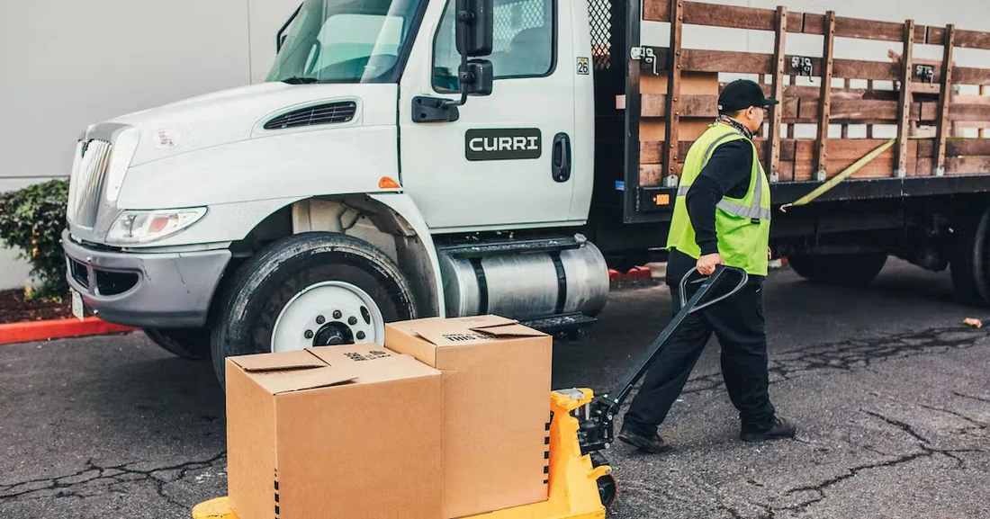 Curri-branded flatbed delivery truck with a driver in safety gear using a pallet jack to move large cardboard boxes off the truck at a commercial delivery site.‍